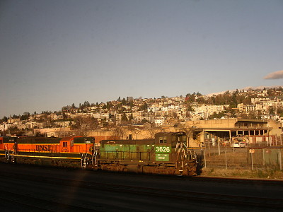 [There is a train engine several tracks over and on the other side of the fence is a hillside with many multi-story white or tan buildings with trees interspersed.]